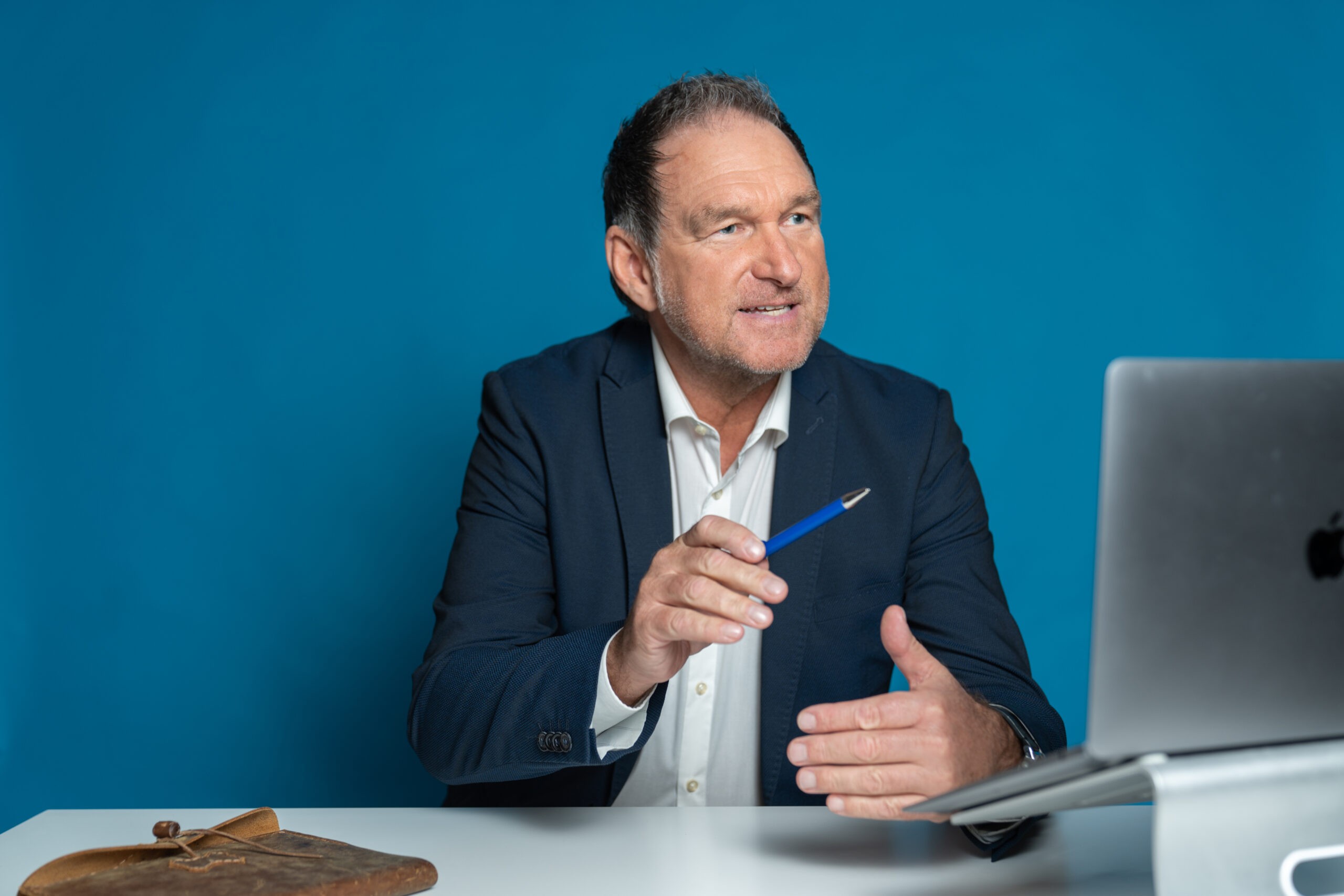 Thomas Gerres, Konferenz-Master, Video Coach sitzt vor einem MacBook und hält einen blauen Kugelschreiber in der Hand